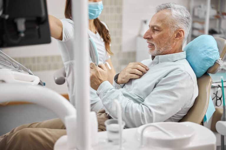 Elderly patient of a dentists office sitting in dental chair and looking in the mirror with a doctor by his side