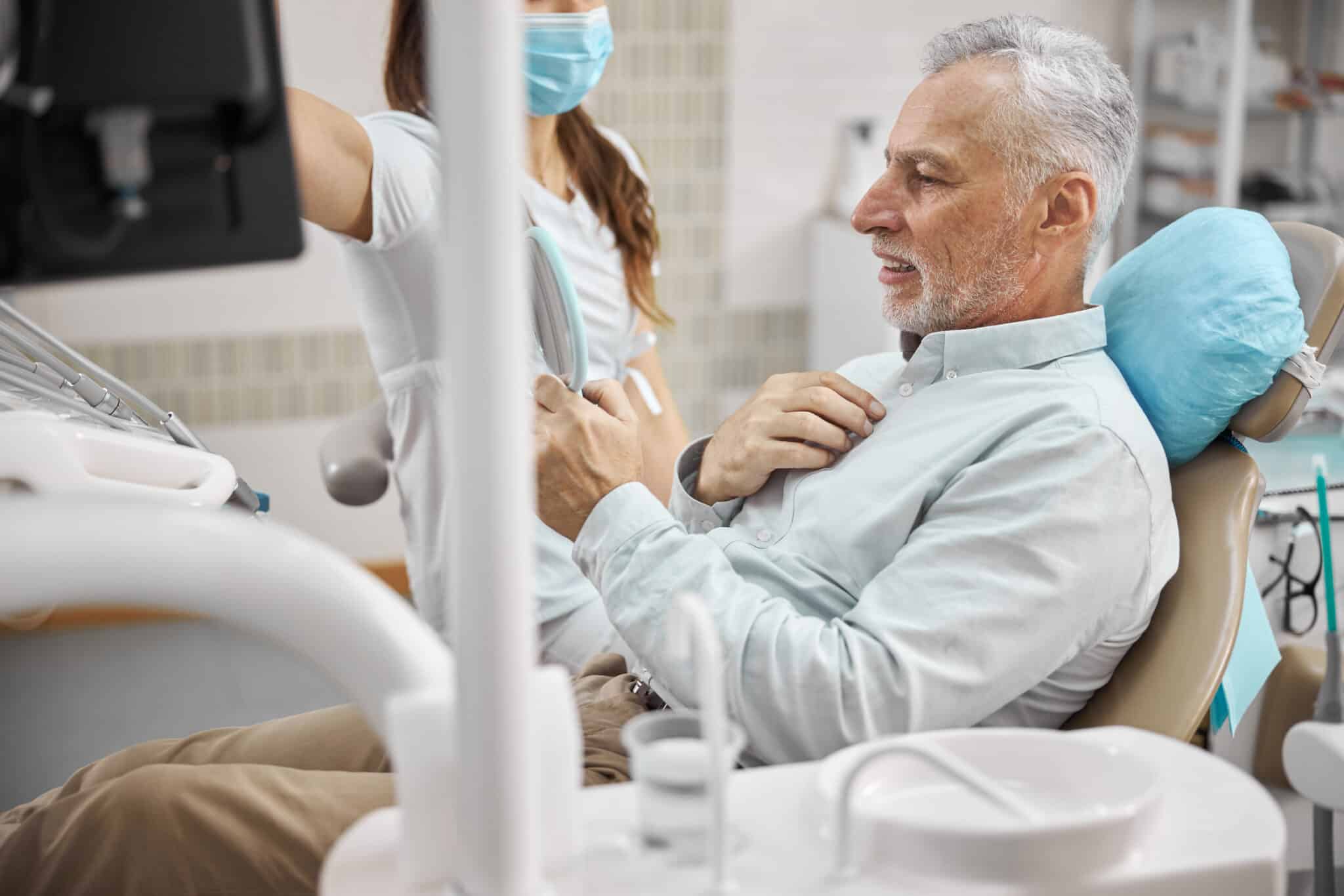 Elderly patient of a dentists office sitting in dental chair and looking in the mirror with a doctor by his side